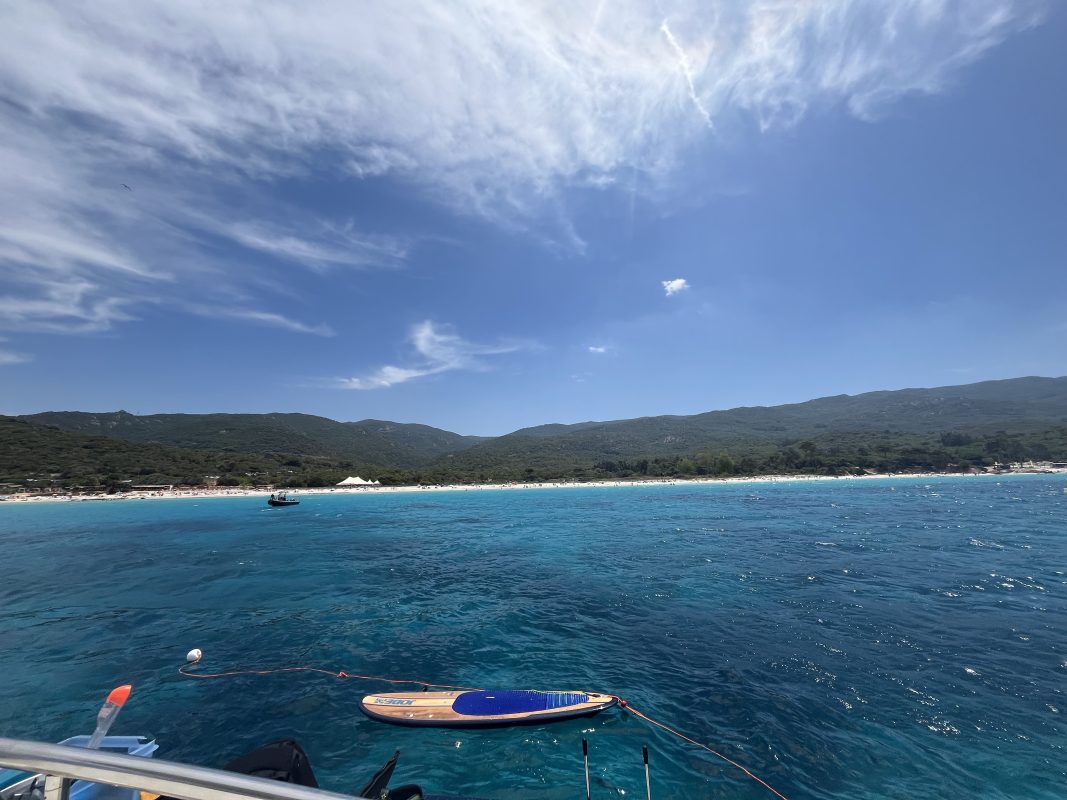 A paddleboard floating beside a yacht in front of a long sandy beach in Corsica