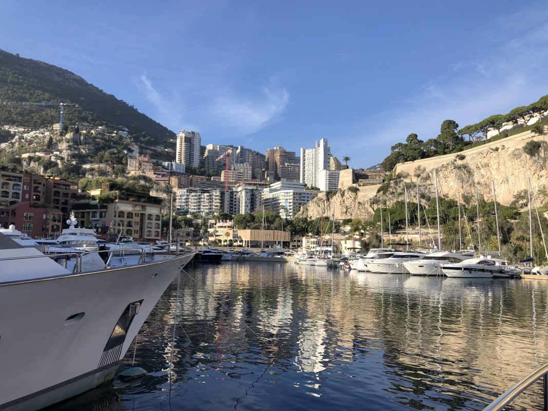 A yacht and moored boats in a Monaco marina with the Rock and city buildings behind
